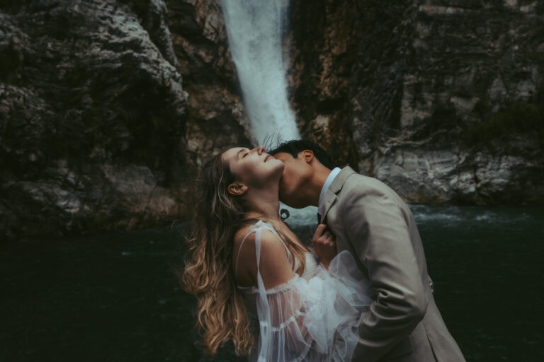 A groom kisses his bride on the neck as she leans back, both of them standing in front of a Swiss waterfall