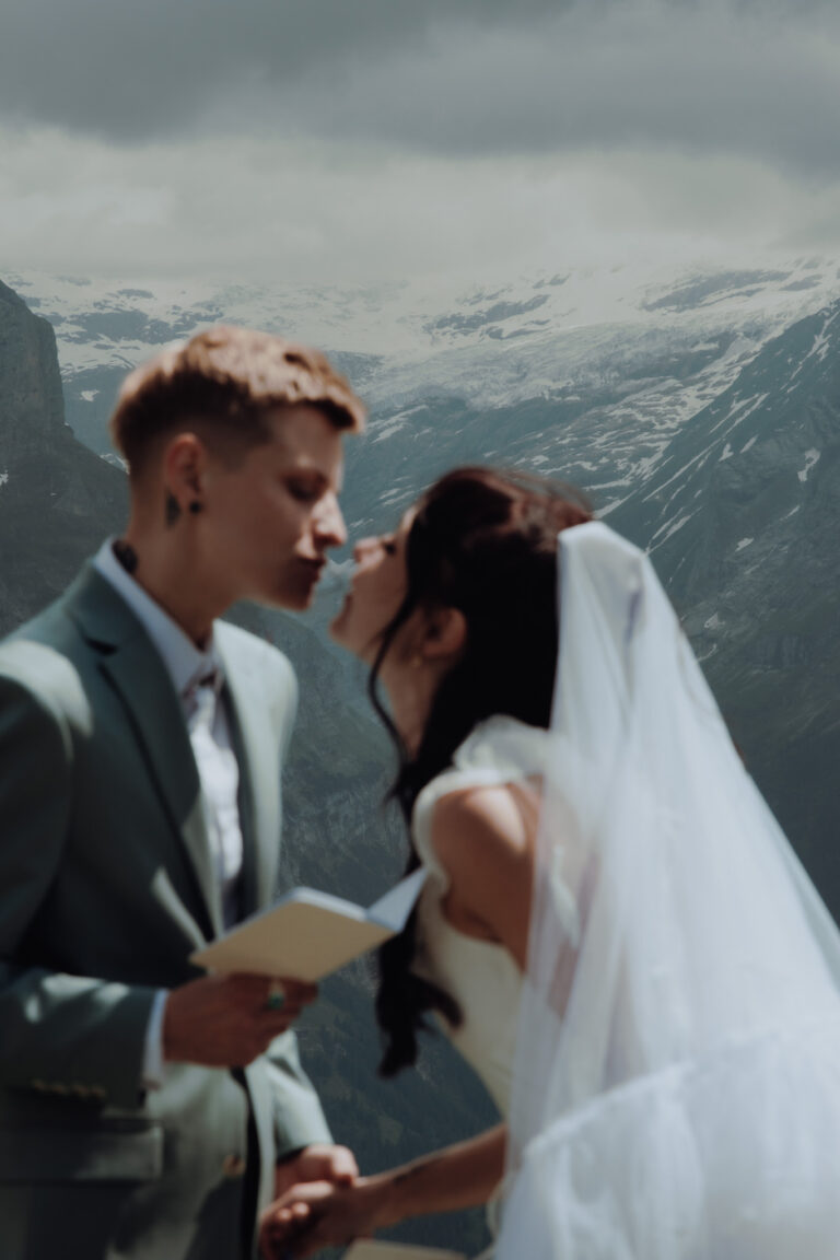 A bride leans forward in the middle of the swiss alps to kiss her bride during their elopement, with the mountains in focus in the background