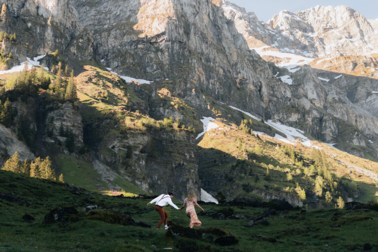 A couple runs together in a field in front of majestic mountain peaks during their engagement session in Switzerland