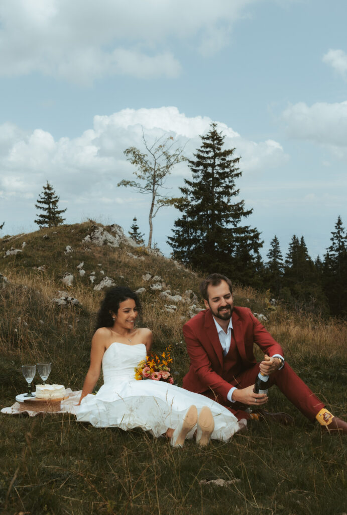During their elopement in the swiss alps, a couple sits on a picnic, and the groom pops a bottle of champagne