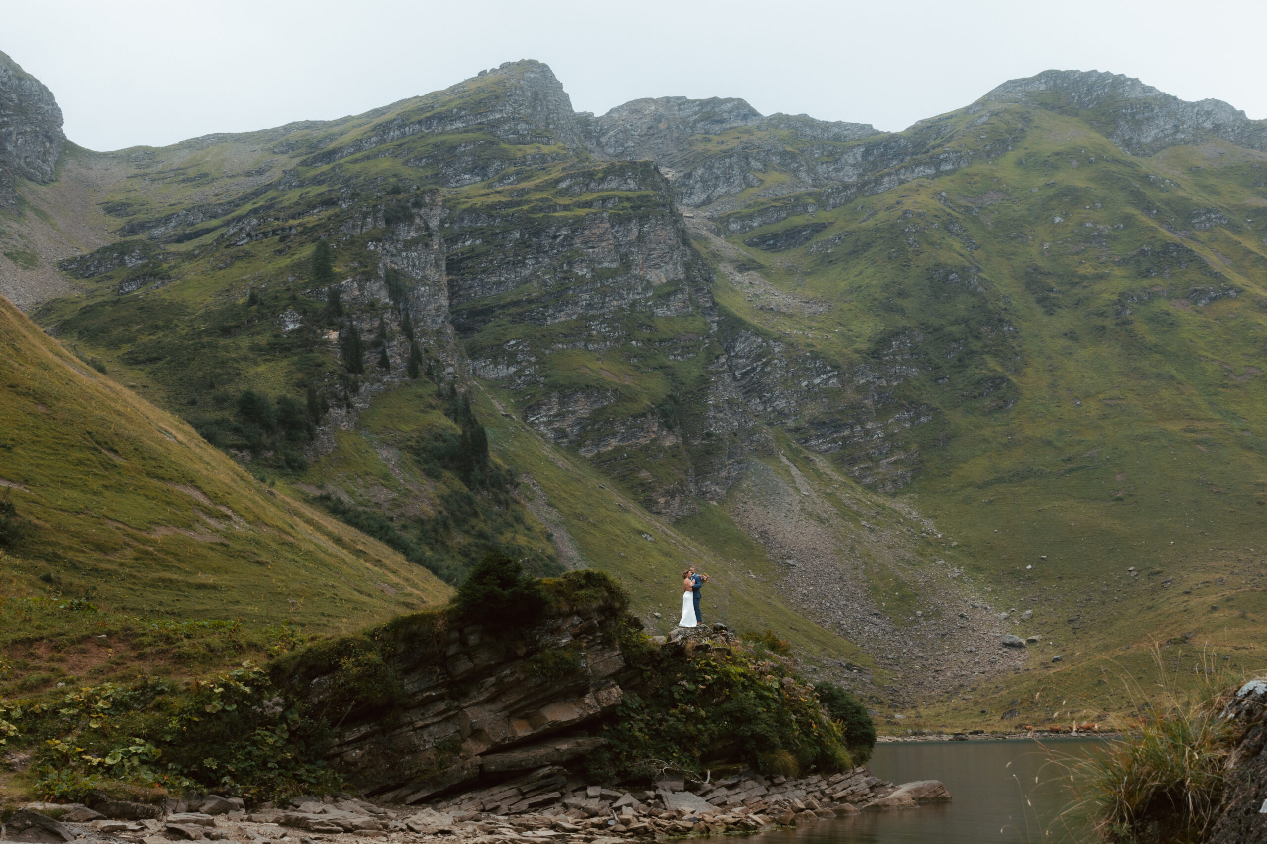 A couple stands on a rock in the distance, in front of an alpine mountain lake, during their elopement in switzerland