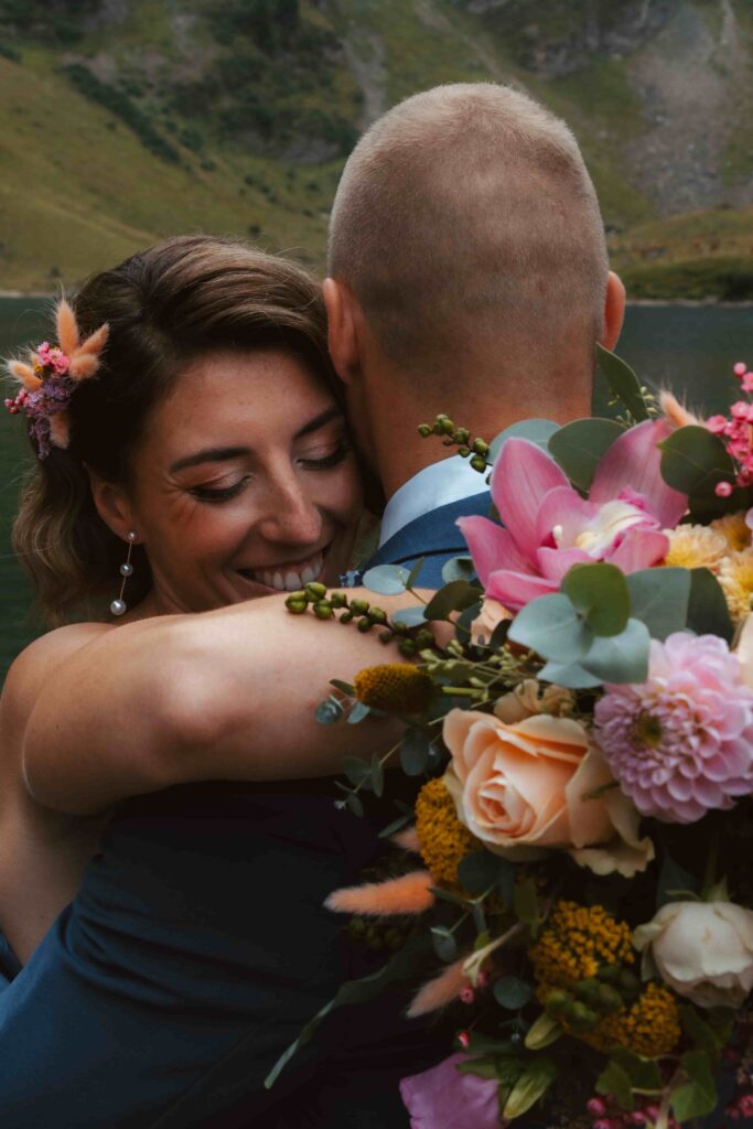 A couple hugs during their elopmenent at an alpin e lake in switzerland. The bride holds a colorful bouquet in her hand